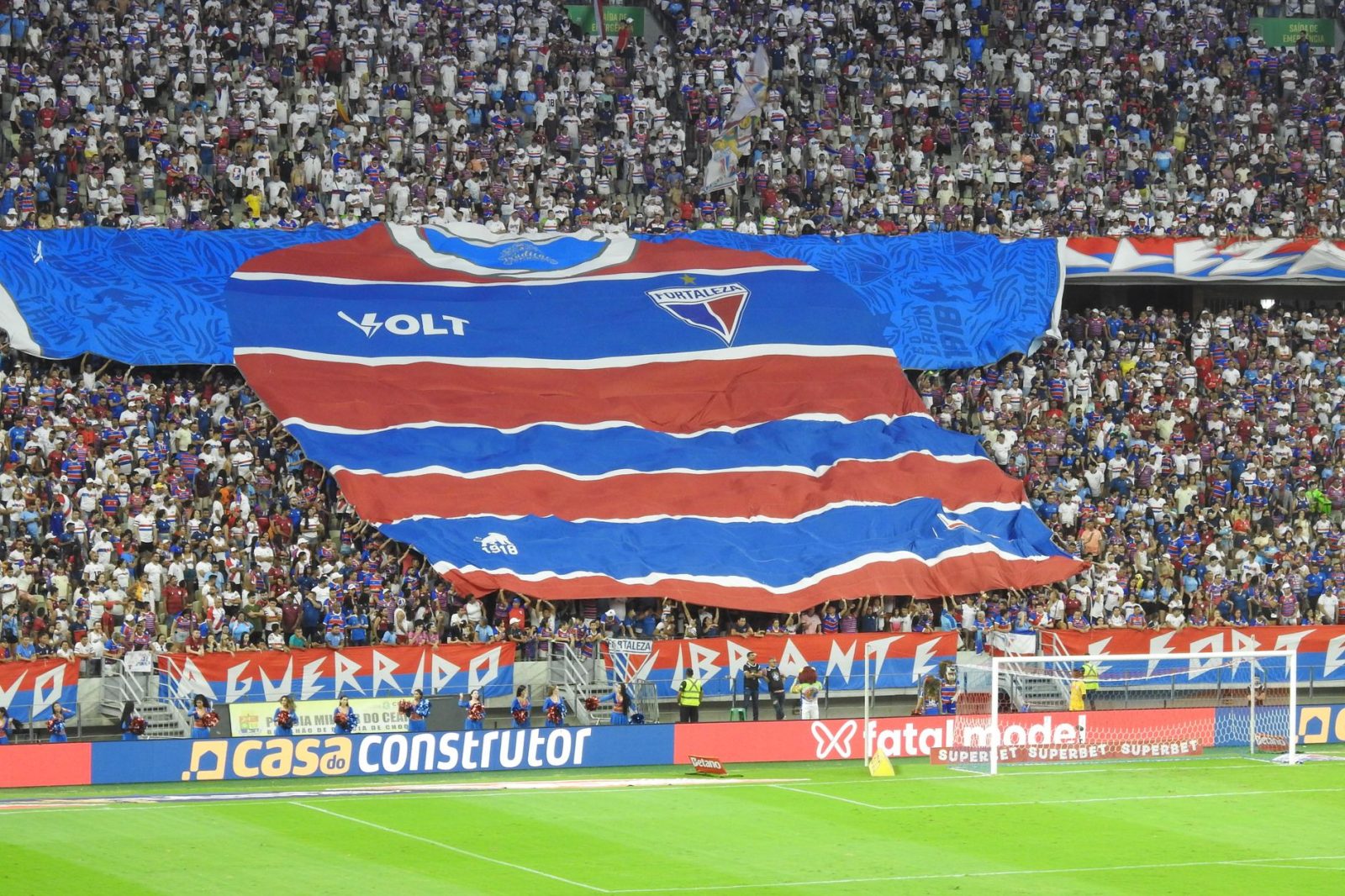Torcedores exibem uma grande faixa com a camisa do Fortaleza nas arquibancadas de um estádio lotado.