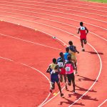Atletas disputam uma corrida em uma pista curva ao ar livre.