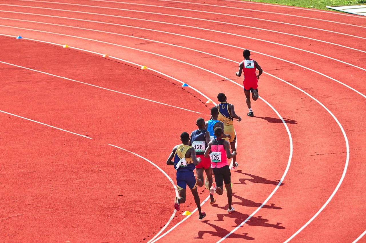 Atletas disputam uma corrida em uma pista curva ao ar livre.