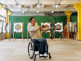 Atleta em cadeira de rodas pratica tiro com arco em uma pista indoor.