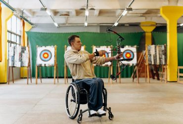 Atleta em cadeira de rodas pratica tiro com arco em uma pista indoor.