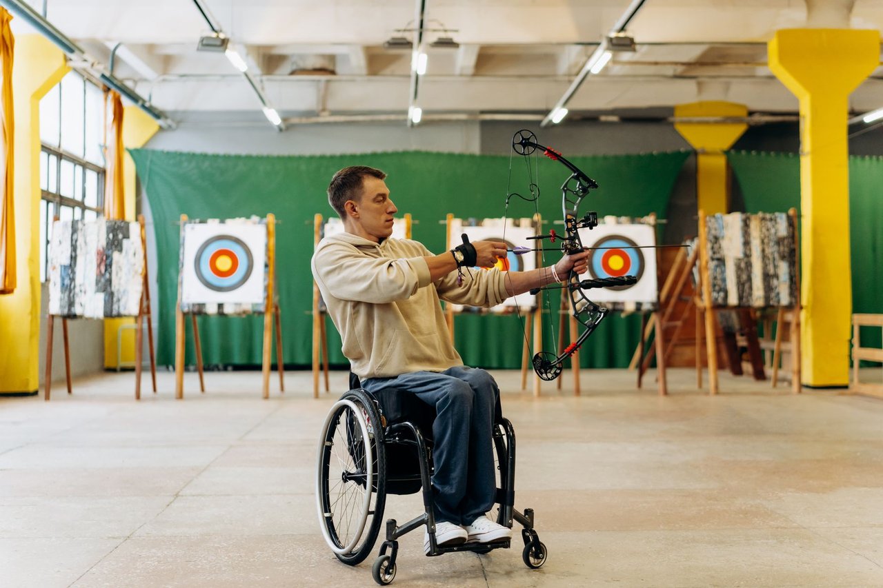 Atleta em cadeira de rodas pratica tiro com arco em uma pista indoor.